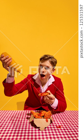 Man holding crispy chicken while pouring mustard sauce with exaggerated facial expression against bold yellow background. Man holding crispy chicken while pouring mustard sauce with exaggerated facial expression against bold yellow background. 125331597