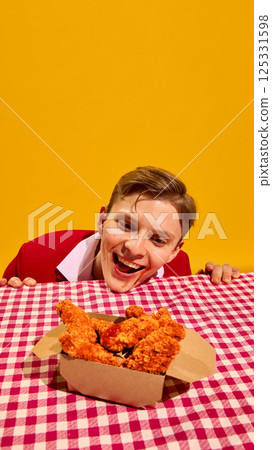 Man leaning over table smiling with excitement looking at box with crispy chicken nuggets against yellow backdrop. Man leaning over table smiling with excitement looking at box with crispy chicken nuggets against yellow backdrop. 125331598