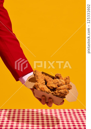 Hand in red sleeve holding box of crispy fried chicken nuggets against yellow background over checkered tablecloth. Hand in red sleeve holding box of crispy fried chicken nuggets against yellow background over checkered tablecloth. 125331602