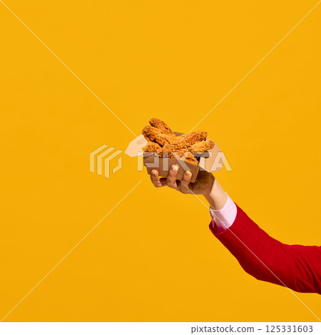 Raised hand holding crispy chicken nuggets box against yellow bold background. 125331603