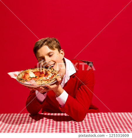 Man with closed eyes and delightful face holding plate with pizza a over red tablecloth against bold red background. 125331609
