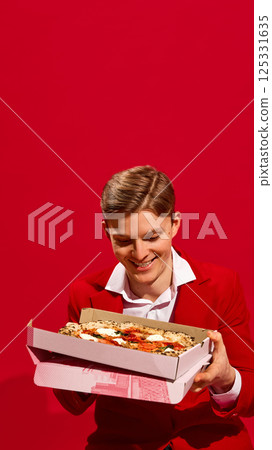 Smiling handsome man in red suit holding pizza box with excitement against vivid red background. 125331635