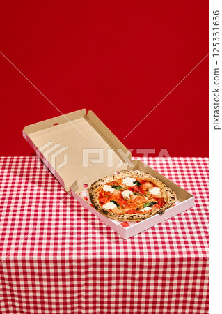 Open pizza box with delicious pizza on red checkered tablecloth against bold red background. 125331636