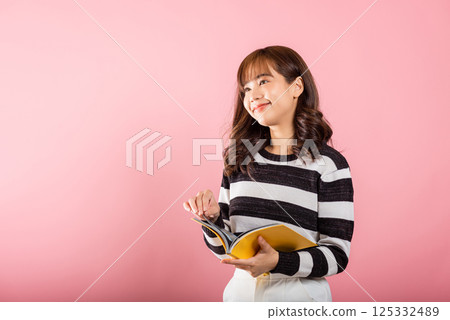 Portrait of beautiful Asian young woman teenage smiling holding a yellow book and reading, studio shot isolated on pink background with copy space 125332489