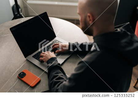 View from shoulder of focused bald programmer with beard and hoodie sitting at table in home office, concentrating on work on laptop computer looking to screen, mobile phone nearby. 125333252