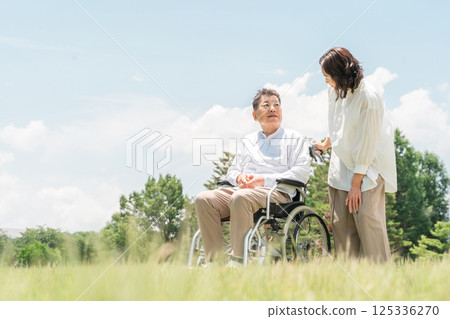 A senior man in a wheelchair in a park and a woman assisting him (caregiving, parent-child, elderly care home) 125336270