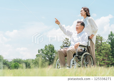 A senior man in a wheelchair in a park and a woman assisting him (caregiving, parent-child, elderly care home) 125336271