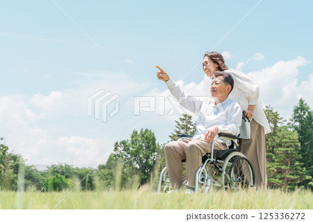 A senior man in a wheelchair in a park and a woman assisting him (caregiving, parent-child, elderly care home) 125336272