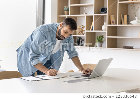 Focused millennial man stand by workplace browse internet write notes Focused millennial man stand by workplace browse internet write notes 125336548