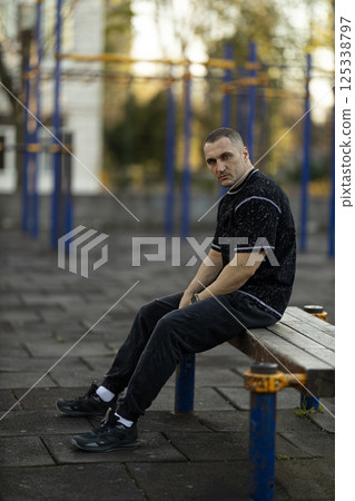 man in casual athletic wear sits on wooden bench in well maintained sports area during afternoon. He appears reflective and focused on his fitness journey. 125338797