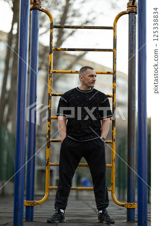 man wearing casual workout clothes stands confidently outdoor fitness park. He is positioned near exercise equipment surrounded urban scenery and greenery. man wearing casual workout clothes stands confidently outdoor fitness park. He is positioned near exercise equipment surrounded urban scenery and greenery. 125338834
