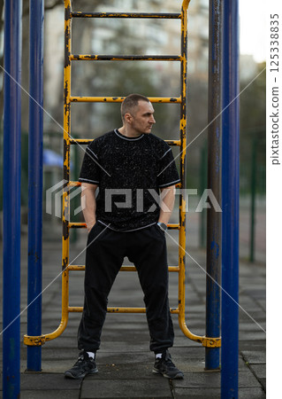 man wearing black sporty outfit is standing yellow exercise equipment in urban park. It is morning and area is quiet greenery visible in background. man wearing black sporty outfit is standing yellow exercise equipment in urban park. It is morning and area is quiet greenery visible in background. 125338835