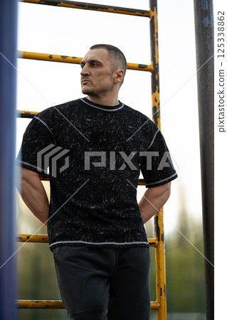 man stands near climbing frames in playground wearing casual black shirt and dark pants. soft sunlight enhances scene as he gazes thoughtfully into distance. 125338862