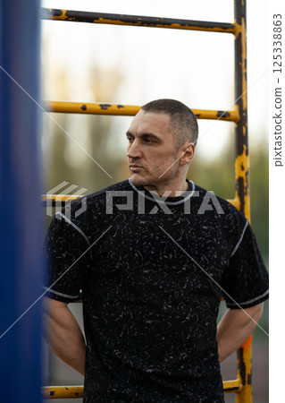 male athlete stands confidently set of exercise bars dressed in fitness attire. golden hour light casts warm glow highlighting his focused expression and strong physique. 125338863