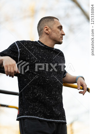 man is focused and relaxed while leaning against exercise equipment in park. sun is setting casting warm light enhancing fitness atmosphere around him. 125338864