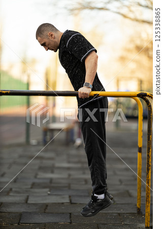 man in athletic wear executes dips on parallel bars while exercising in public park during late afternoon. setting features trees and playground equipment in background. 125338865