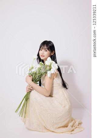 Portrait of a young woman sitting wearing a white dress and holding a bouquet of white flowers 125339211