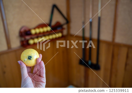 Hand holding billiard ball number 12 in front of a home billiard setup with cues and rack. Concept of indoor recreation and personal hobby space Hand holding billiard ball number 12 in front of a home billiard setup with cues and rack. Concept of indoor recreation and personal hobby space 125339911