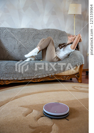 Robot vacuum cleaning the floor while a young woman relaxes on the couch in the background. Concept of smart home technology and effortless cleaning 125339964