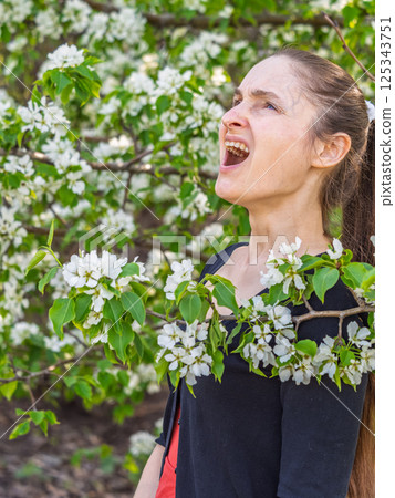 Young woman sneezes in the park against the background of a flowering tree. Allergy to pollen concept. Young woman sneezes in the park against the background of a flowering tree. Allergy to pollen concept. 125343751