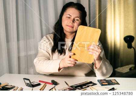 Woman looking in mirror before doing makeup. Table with decorative cosmetics. Preparation for the event. Beauty and self-care concept. 125348780