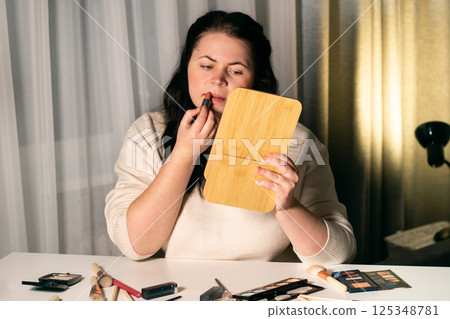 Woman looking in mirror and applying lipstick to lips. Self makeup. Preparing for event. Beauty and self-care concept 125348781