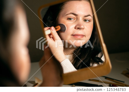 Woman doing make up looking in mirror at home. Putting on make-up with brush. Applying skin powder on face. Covering red spots. Reflection in mirror. 125348783