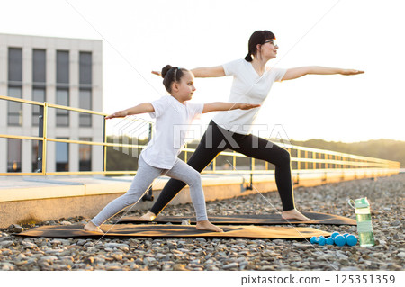 Mother and daughter, adult female and young girl, performing yoga poses on rooftop. Fitness, balance, and bonding activity amid open sky offering family moments of wellness and tranquility. 125351359