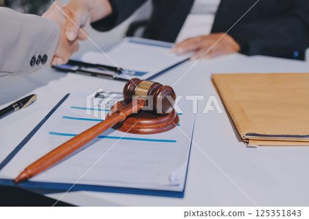 Two businesspeople are shaking hands in a law office with a scale and a gavel on the table 125351843