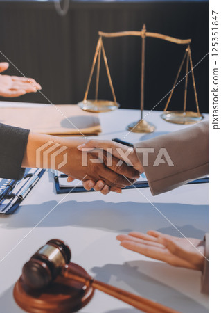 Two businesspeople are shaking hands in a law office with a scale and a gavel on the table 125351847