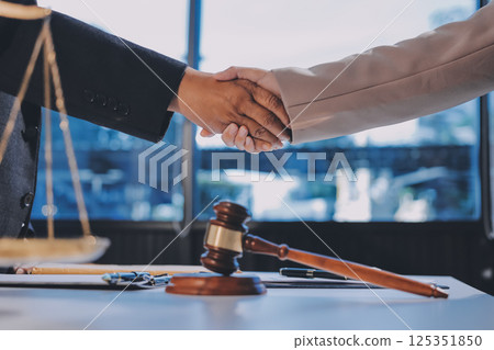 Two businesspeople are shaking hands in a law office with a scale and a gavel on the table 125351850