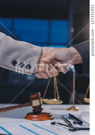 Two businesspeople are shaking hands in a law office with a scale and a gavel on the table Two businesspeople are shaking hands in a law office with a scale and a gavel on the table 125351852