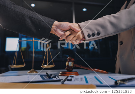 Two businesspeople are shaking hands in a law office with a scale and a gavel on the table 125351853