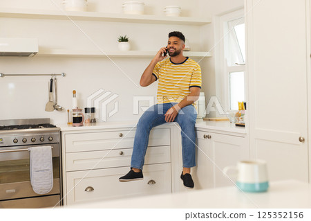 Young man sitting on kitchen counter, talking on smartphone, enjoying conversation Young man sitting on kitchen counter, talking on smartphone, enjoying conversation 125352156