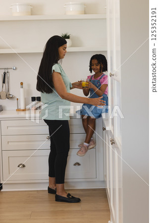 Mother giving juice to daughter sitting on kitchen counter, both smiling warmly Mother giving juice to daughter sitting on kitchen counter, both smiling warmly 125352191