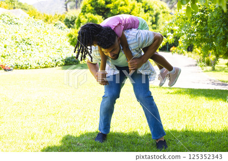 Father giving daughter piggyback ride in sunny park, both laughing together 125352343