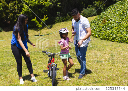 Father teaching daughter to ride bicycle while mother supports in sunny park 125352344