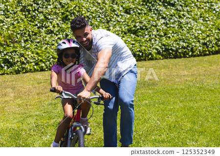 Father teaching daughter to ride bicycle in park, both smiling and enjoying time 125352349