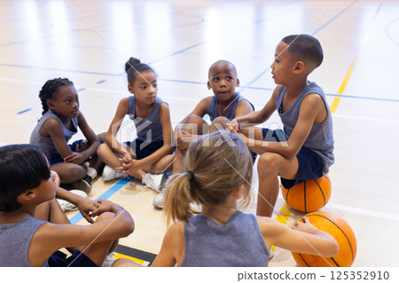 In school gym, multiracial children sitting on floor listening to teammate, holding basketball 125352910