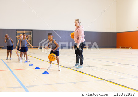 In school gym, diverse children practicing basketball drills with female coach supervising In school gym, diverse children practicing basketball drills with female coach supervising 125352938