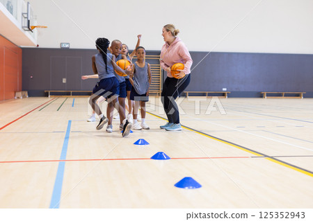 Playing basketball, diverse children in school gym practicing with female coach and cones Playing basketball, diverse children in school gym practicing with female coach and cones 125352943