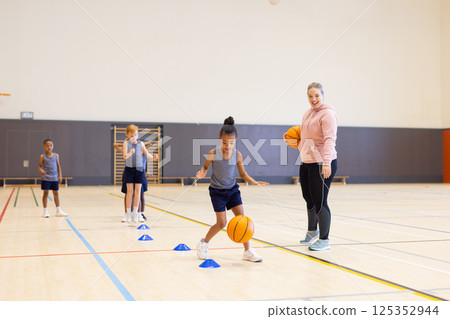 In school gym, diverse children practicing basketball drills with female coach 125352944