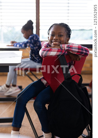 Smiling african american girl sitting on chair with backpack in school classroom, enjoying break Smiling african american girl sitting on chair with backpack in school classroom, enjoying break 125352955