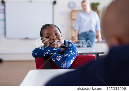 Smiling african american girl in school classroom leaning on chair, female teacher in background 125353006