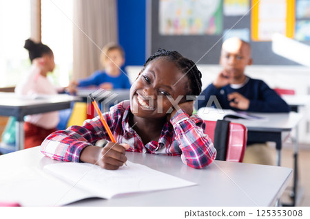 In school, smiling african american girl holding pencil and writing in notebook in classroom 125353008