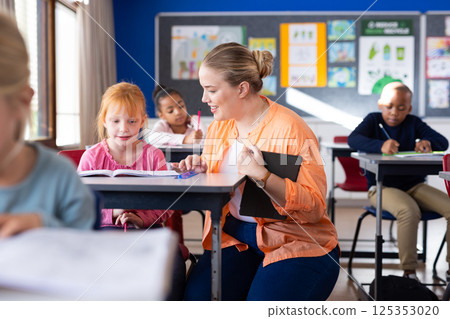 In school, female teacher helping young girl with reading at desk in classroom 125353020