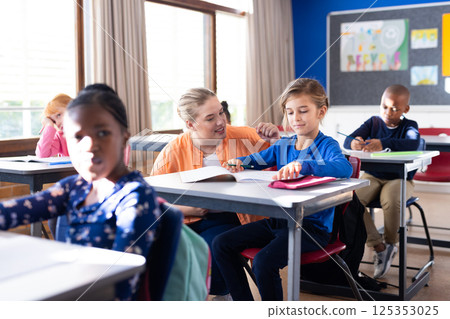 Helping boy with homework, female teacher in school classroom, other students studying 125353025