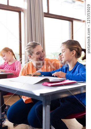 In school, female teacher smiling and helping boy with reading at desk 125353029