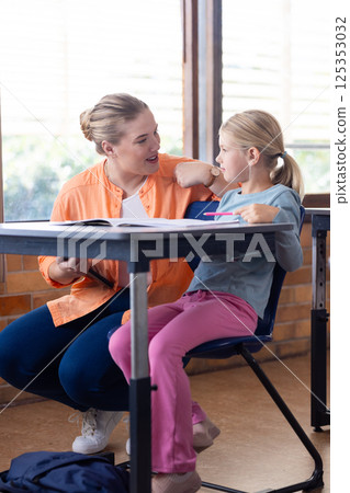 Female teacher helping young girl with schoolwork in classroom, engaging in discussion 125353032
