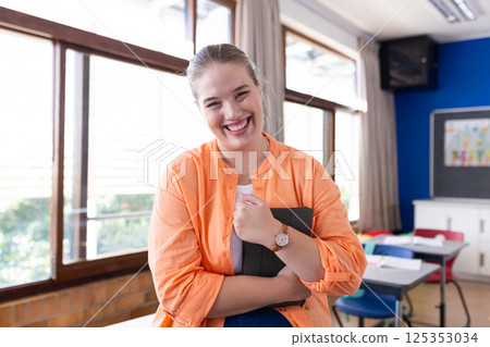Smiling female teacher holding tablet in classroom, standing near desks in school 125353034